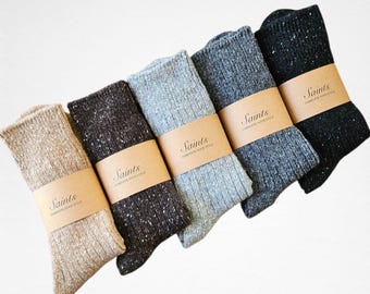 Op de afbeelding: Vijf paar sokken van getextureerd breiwerk in verschillende tinten beige, bruin, grijs en zwart. Elke paar is verpakt met een bruine papieren band met de tekst "Saints COMPLETE YOUR STYLE". De sokken zijn diagonaal geplaatst tegen een witte achtergrond.