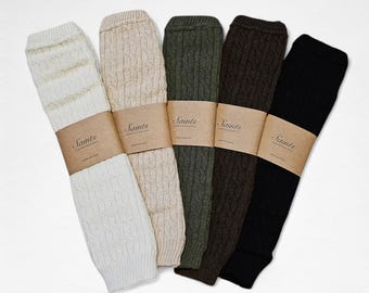 May include: Five cable-knit leg warmers in cream, beige, olive green, dark brown, and black. Each pair is wrapped with a brown paper band that reads "Saints". The leg warmers are arranged in a fan shape against a white background.