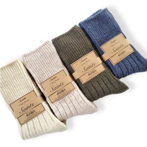 K&ouml;nnte beinhalten: Vier Paar Socken in verschiedenen Farben: Beige, Olivgr&uuml;n, Blau und Wei&szlig;. Die Socken sind aus Lammwolle und haben eine gerippte Struktur. Jedes Paar hat ein braunes Etikett mit dem aufgedruckten Text "Saints Lamb's Wool Saints".