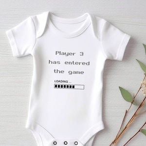 May include: White baby bodysuit with the text "Player 3 has entered the game" and a loading bar graphic in black. The bodysuit is short-sleeved and has poppers at the bottom. A sprig of dried flowers with green leaves is next to the bodysuit.
