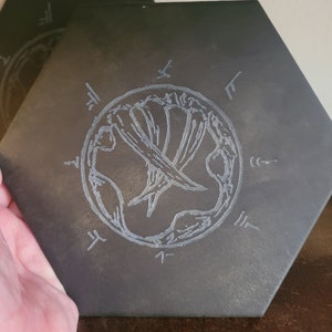 Könnte beinhalten: Ein schwarzer sechseckiger Schieferuntersetzer mit einem weißen, geätzten Design eines stilisierten Baumes, der von einem Kreis umgeben ist. Der Baum hat ein Symbol aus zwei gekreuzten Schwertern in der Mitte. Der Kreis ist mit Runen umrandet.