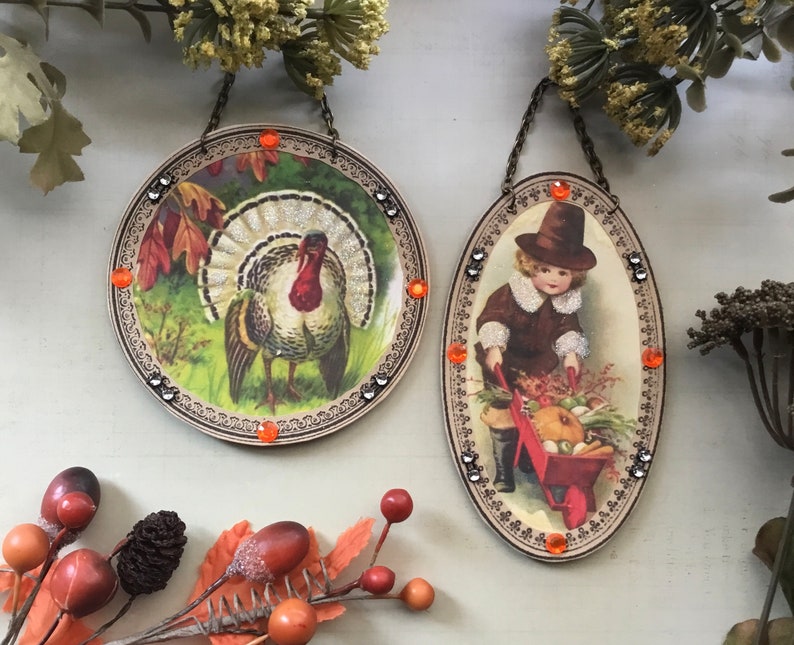 May include: Two round, decorative, framed ornaments with orange and silver embellishments. The ornament on the left features a colorful turkey illustration. The ornament on the right features a child in a brown coat and hat pushing a red wagon full of vegetables.