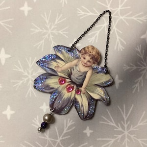 May include: A purple and white flower ornament featuring a girl in a blue dress. The ornament has pink glitter accents and hangs from a silver chain with a pearl and purple bead drop.