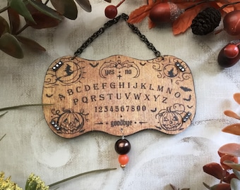 May include: A wooden Halloween-themed Ouija board with a black chain, a brown and orange pendulum, and the words "yes", "no", and "goodbye" printed on it.