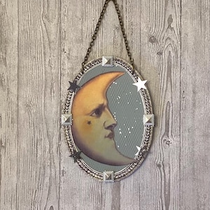 May include: A decorative oval mirror with a silver frame and a crescent moon with a face inside. The moon is surrounded by silver stars and rhinestones.