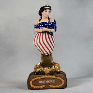 Puede incluir: Una figurita de porcelana de una mujer vestida con un vestido de bandera estadounidense rojo, blanco y azul. La figurita está de pie sobre una base dorada con una base de madera que dice "BENNMORE".