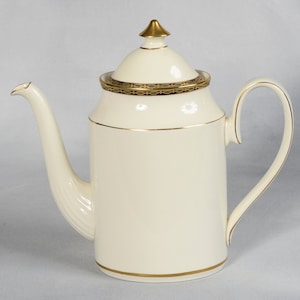 May include: A white ceramic coffee pot with a gold trim and a gold lid. The pot has a rounded body and a spout on the side. The handle is attached to the side of the pot.