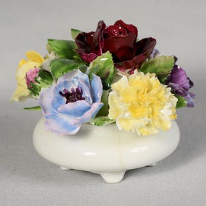 Może przedstawiać: Ceramiczna kompozycja kwiatowa z głęboką czerwoną różą, żółtymi goździkami oraz niebieskimi i fioletowymi kwiatami. Kwiaty ułożone są w małym, białym wazonie na nóżkach. Dekoracyjny element wystroju wnętrz.