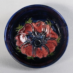 May include: A blue ceramic bowl with a raised, three-dimensional design of a red flower with a dark center. The bowl is glazed with a deep blue color.