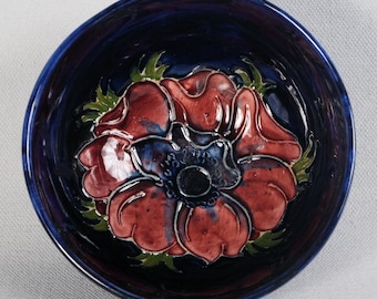 May include: A blue ceramic bowl featuring a raised, three-dimensional design of a red flower with a dark centre. The bowl is glazed in a deep blue colour.