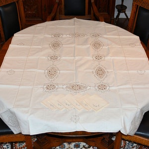 Puede incluir: Un mantel blanco bordado con un delicado patrón floral y cinco servilletas a juego. El mantel está puesto sobre una mesa redonda de madera con sillas negras.