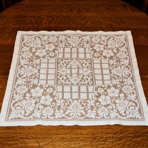 Puede incluir: Un tapete cuadrado de encaje blanco roto con un diseño floral y geométrico. El tapete está colocado sobre una mesa de madera oscura. El patrón de encaje presenta flores y un centro en forma de rejilla, creando una estética vintage.