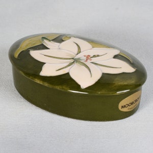 Pode incluir: Uma caixa de cerâmica oval com um esmalte verde e um desenho de flor de lírio branco. A caixa tem um rótulo dourado na parte inferior que diz "Moorcroft England".
