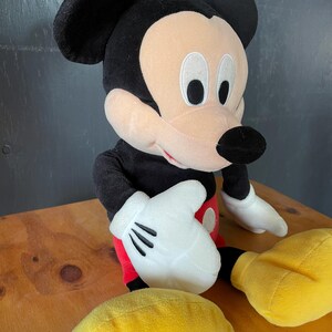 Op de afbeelding: Een pluche Mickey Mouse speelgoed met zwarte vacht, een rode broek en gele schoenen. Het speelgoed heeft een grote glimlach en grote, zwarte ogen.