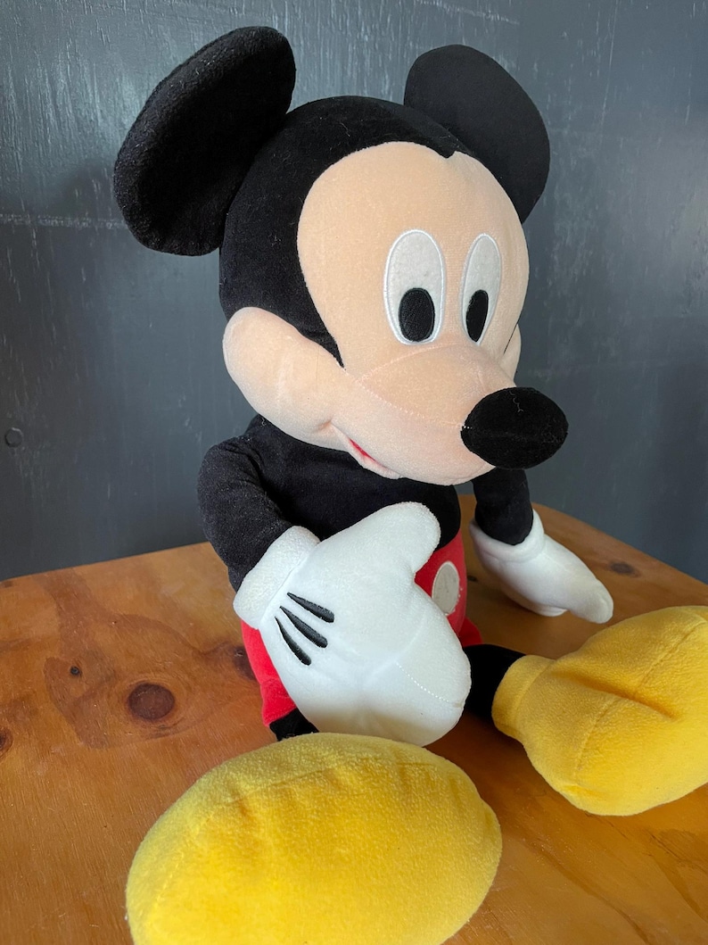 Op de afbeelding: Een pluche Mickey Mouse speelgoed met zwarte vacht, een rode broek en gele schoenen. Het speelgoed heeft een grote glimlach en grote, zwarte ogen.