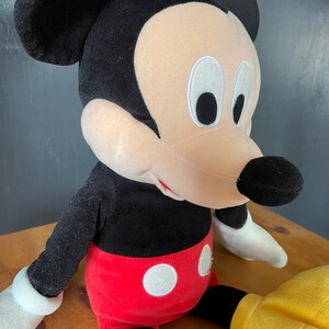 Op de afbeelding: Een pluche Mickey Mouse speelgoed met zwarte vacht, rode shorts met witte stippen en grote witte handschoenen. Het speelgoed heeft een vriendelijke uitdrukking met zwarte ogen en een brede glimlach.