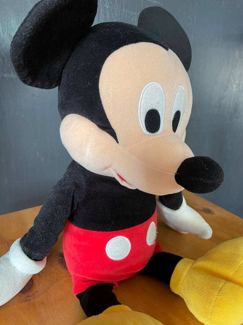 Op de afbeelding: Een pluche Mickey Mouse speelgoed met zwarte vacht, rode shorts met witte stippen en grote witte handschoenen. Het speelgoed heeft een vriendelijke uitdrukking met zwarte ogen en een brede glimlach.