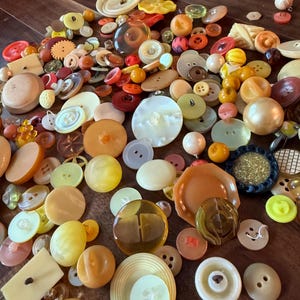 May include: A large collection of assorted buttons in various shades of yellow, orange, cream, and brown. The buttons vary in size, shape, and material, including round, square, and textured designs, some with multiple holes and others with shanks.