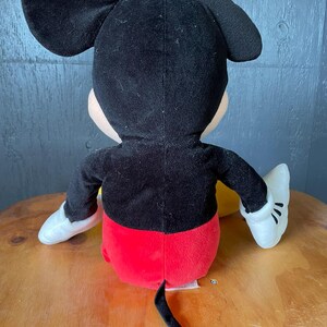 Op de afbeelding: Een pluche Mickey Mouse speelgoed in zwart en rood, zittend op een houten oppervlak. Het speelgoed heeft witte handschoenen en een zwarte staart.