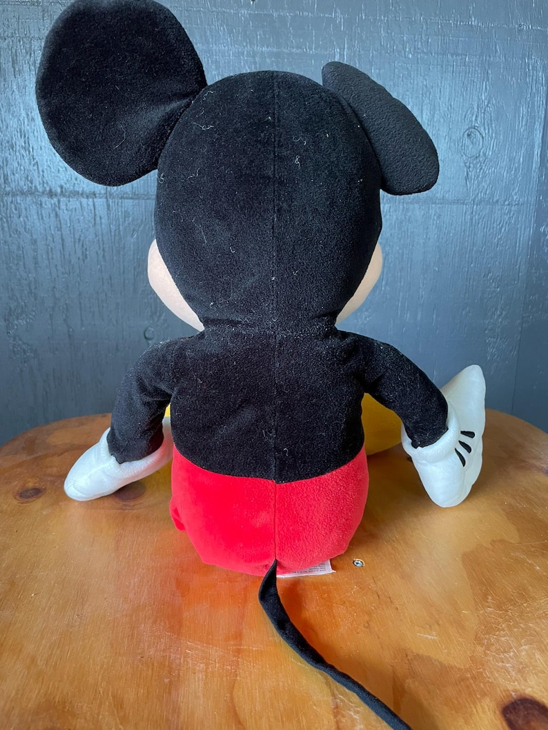 Op de afbeelding: Een pluche Mickey Mouse speelgoed in zwart en rood, zittend op een houten oppervlak. Het speelgoed heeft witte handschoenen en een zwarte staart.