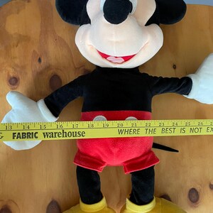 Op de afbeelding: Een groot pluche Mickey Mouse speelgoed met zwarte oren, een zwart lichaam, een rode broek en gele schoenen. Het speelgoed zit op een houten oppervlak met een gele meetlint waarop "FABRIC warehouse WHERE THE BEST IS NOT EX" staat.