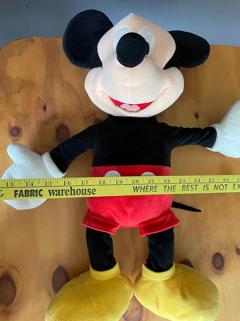 Op de afbeelding: Een groot pluche Mickey Mouse speelgoed met zwarte oren, een zwart lichaam, een rode broek en gele schoenen. Het speelgoed zit op een houten oppervlak met een gele meetlint waarop "FABRIC warehouse WHERE THE BEST IS NOT EX" staat.