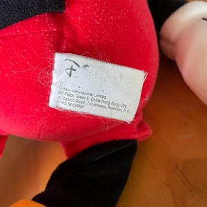 Op de afbeelding: Rood en zwart pluche Mickey Mouse speelgoed met een gele en oranje accent. Het speelgoed heeft een label met de tekst "Mean International Limited. 8th Floor, Tower 5, China Hong Kong City, 33 Canton Road, Tsimshatsui, Kowloon, H.K. MADE IN CHINA"