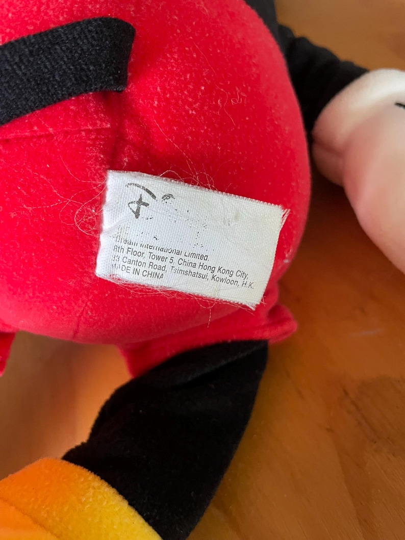 Op de afbeelding: Rood en zwart pluche Mickey Mouse speelgoed met een gele en oranje accent. Het speelgoed heeft een label met de tekst "Mean International Limited. 8th Floor, Tower 5, China Hong Kong City, 33 Canton Road, Tsimshatsui, Kowloon, H.K. MADE IN CHINA"