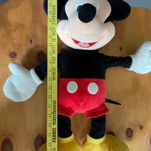 Op de afbeelding: Een groot pluche Mickey Mouse speelgoed met zwarte vacht, een rode broek, gele schoenen en een wit gezicht met zwarte ogen en een rode mond. Het speelgoed is ongeveer 71 cm hoog.