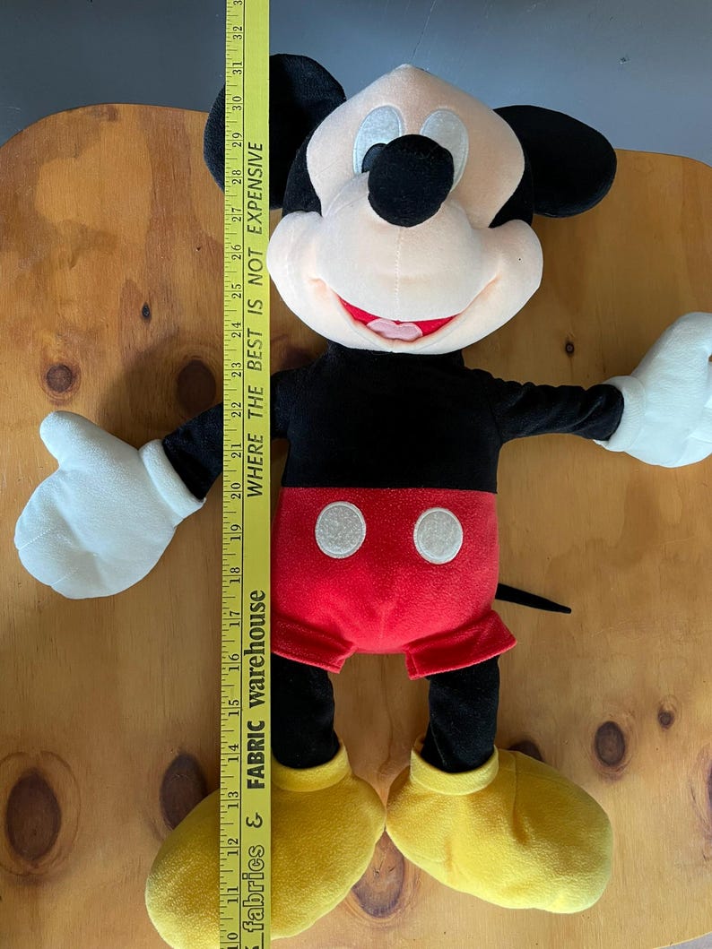 Op de afbeelding: Een groot pluche Mickey Mouse speelgoed met zwarte vacht, een rode broek, gele schoenen en een wit gezicht met zwarte ogen en een rode mond. Het speelgoed is ongeveer 71 cm hoog.