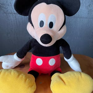 Op de afbeelding: Een groot pluche Mickey Mouse speelgoed dat op een houten oppervlak zit. Mickey draagt zijn kenmerkende rode broek met twee witte knopen en gele schoenen. Hij heeft zwarte oren, een zwarte neus en witte ogen.
