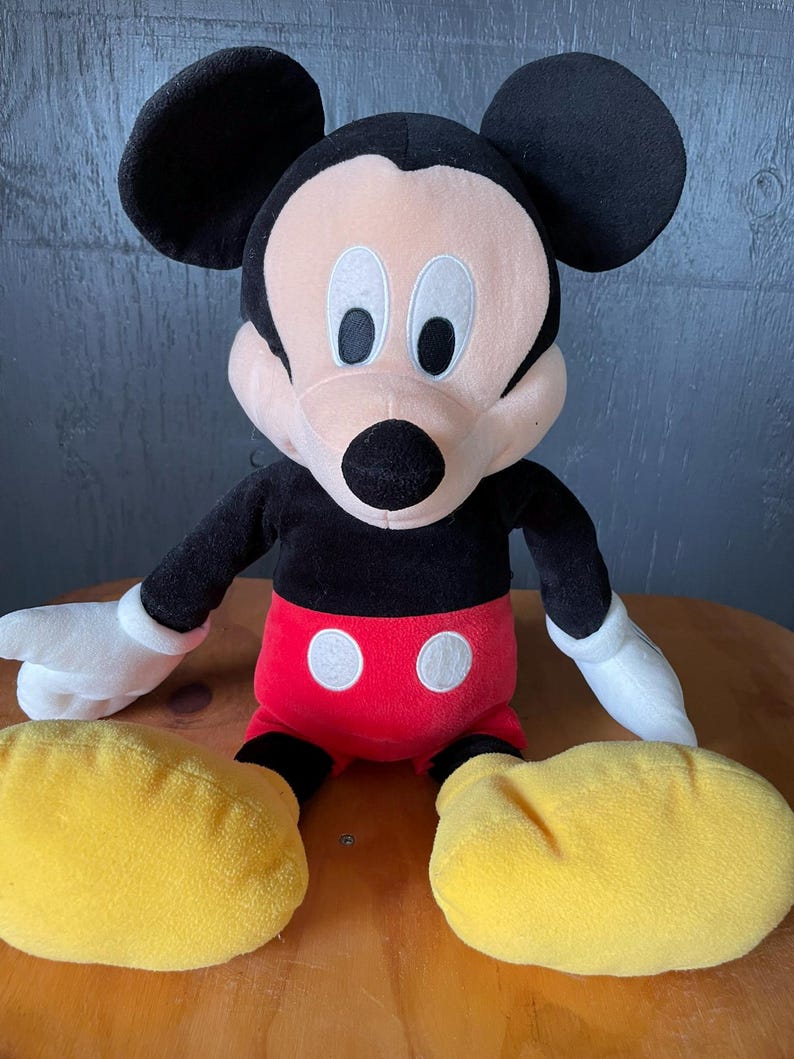 Op de afbeelding: Een groot pluche Mickey Mouse speelgoed dat op een houten oppervlak zit. Mickey draagt zijn kenmerkende rode broek met twee witte knopen en gele schoenen. Hij heeft zwarte oren, een zwarte neus en witte ogen.