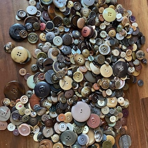 Puede incluir: Un gran surtido de botones vintage en varios tamaños, formas y colores. Los botones están esparcidos sobre una superficie de madera, con tonos que van desde el crema y el beige hasta el marrón y el negro. Muchos tienen cuatro agujeros, otros dos o son botones de caña.