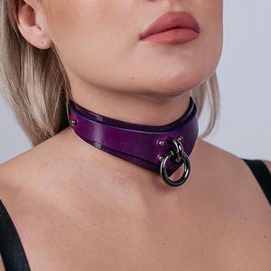 Puede incluir: Un collarín de cuero morado con un anillo y remaches de metal plateado. El collarín se lleva alrededor del cuello y tiene un diseño curvo. La imagen es un primer plano del collarín.