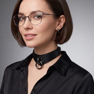 May include: A black leather choker featuring a central metal O-ring attached with two rivets. The choker has a smooth matte finish and is secured with a metal buckle. It is worn with a black collared shirt, silver hoop earrings, and glasses.