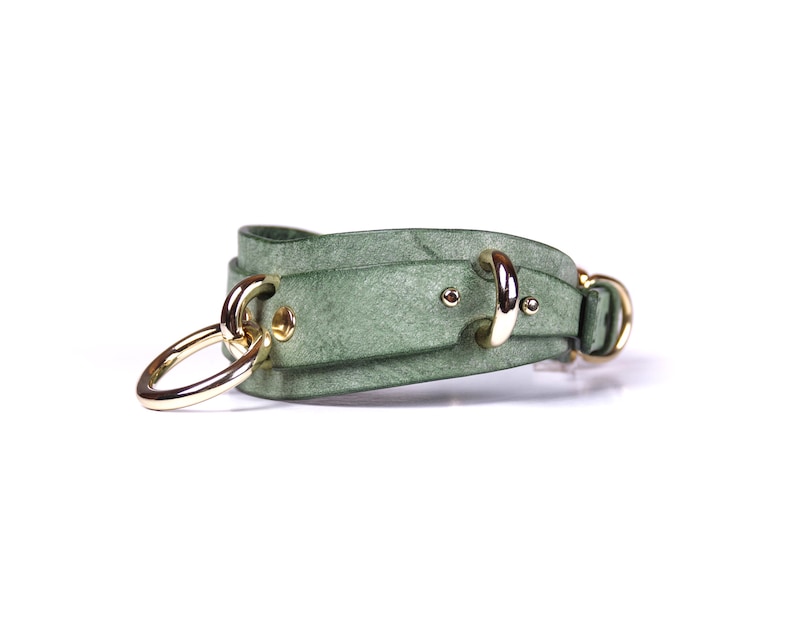 May include: A sage green leather bracelet with gold-colored metal hardware. The bracelet features a double-strap design with two large gold rings and smaller gold accents. The bracelet is shown against a white background.