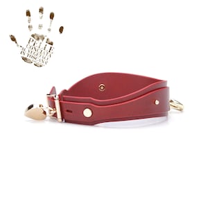 May include: A red leather collar with gold-coloured hardware. The collar has a heart-shaped clasp and a gold ring. The image also includes a handprint with text in Italian.