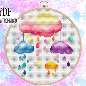 Puede incluir: Un patrón de punto de cruz con un móvil de nubes de colores con nubes rosas, amarillas, azules, moradas y naranjas. Las nubes cuelgan de cuerdas con gotas de lluvia que caen de cada nube.