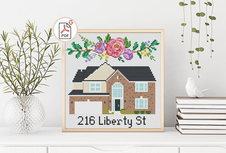 May include: Cross-stitch pattern of a house with a floral wreath above the door. The house is brown brick with a white door and windows. The address "216 Liberty St" is below the house.