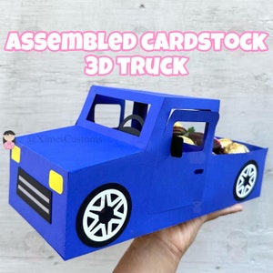 Op de afbeelding: Een blauwe kartonnen 3D-truck met een zwart stuurwiel, zwarte ramen en gele koplampen. De laadbak van de truck is gevuld met chocoladebonbons. De tekst "Assembled Cardstock 3D Truck" staat bovenaan de afbeelding.