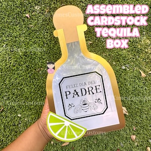 Könnte beinhalten: Zusammengebaute Tequila-Box aus Karton in Form einer Tequila-Flasche. Die Box hat einen goldenen Rand und einen silbernen Innenraum. Das Etikett lautet "Feliz Día del Padre" mit einer Bienenillustration. Ein Limettenschnitz ist enthalten.