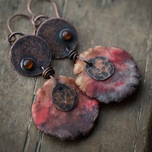 Może przedstawiać: Ręcznie robione kolczyki z miedzi, z okrągłym wzorem z bursztynowymi kamieniami i teksturowanymi, czerwonobrązowymi elementami gliny. Kolczyki mają rustykalną, artystyczną estetykę, z naciskiem na naturalne materiały.