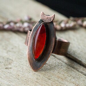 Può includere: Anello in rame fatto a mano con una gemma ovale rosso intenso. L'anello ha un dettaglio a forma di mezzaluna sulla parte superiore. Appoggiato su una superficie di legno.