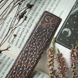 May include: A detailed, dark metal bookmark with a copper-colored leaf and vine design. The top of the bookmark features a crescent moon and stars. The bookmark is resting on an open book with text.