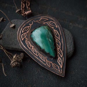 Puede incluir: Colgante de cobre hecho a mano con forma de lágrima. El colgante presenta una piedra verde central de forma irregular. El marco de cobre tiene una pátina oscura y está adornado con un diseño de hojas. Se adjunta una cadena de cobre.