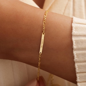 May include: A gold-colored bracelet with a chain and a rectangular bar engraved with the name "CHLOE". The bracelet is displayed on a person's wrist, with a cream-colored sweater in the background.