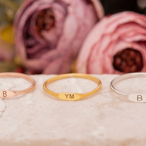 May include: Three delicate rings in rose gold, gold, and silver, each with a small, engraved oval plate. The rose gold ring is engraved with the letter "B", the gold ring with "YM", and the silver ring with "B".