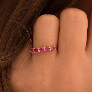 May include: A gold ring featuring a row of vibrant red gemstones, interspersed with small, sparkling white diamonds. The ring is set against a blurred background, highlighting the jewelry's intricate design and color contrast.
