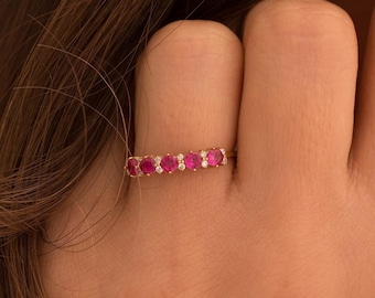 May include: A gold ring featuring a row of vibrant red gemstones, interspersed with small, sparkling white diamonds. The ring is set against a blurred background, highlighting the jewelry's intricate design and color contrast.