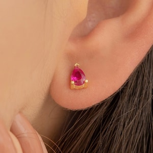 May include: A pair of gold stud earrings with a teardrop-shaped pink gemstone.